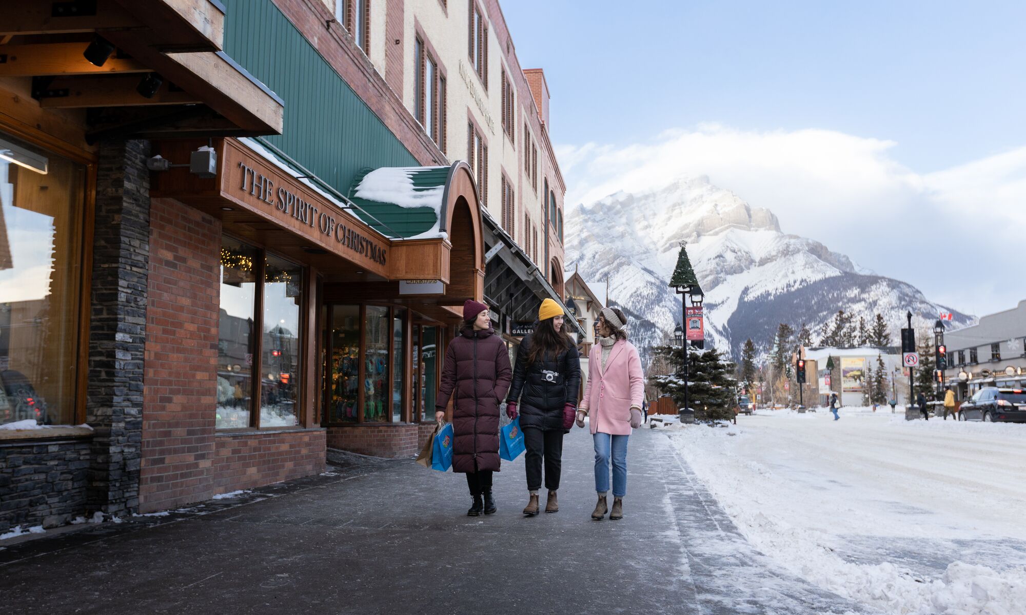 Three girls walk down Banff Ave while shopping with Cascade Mountain in the background.